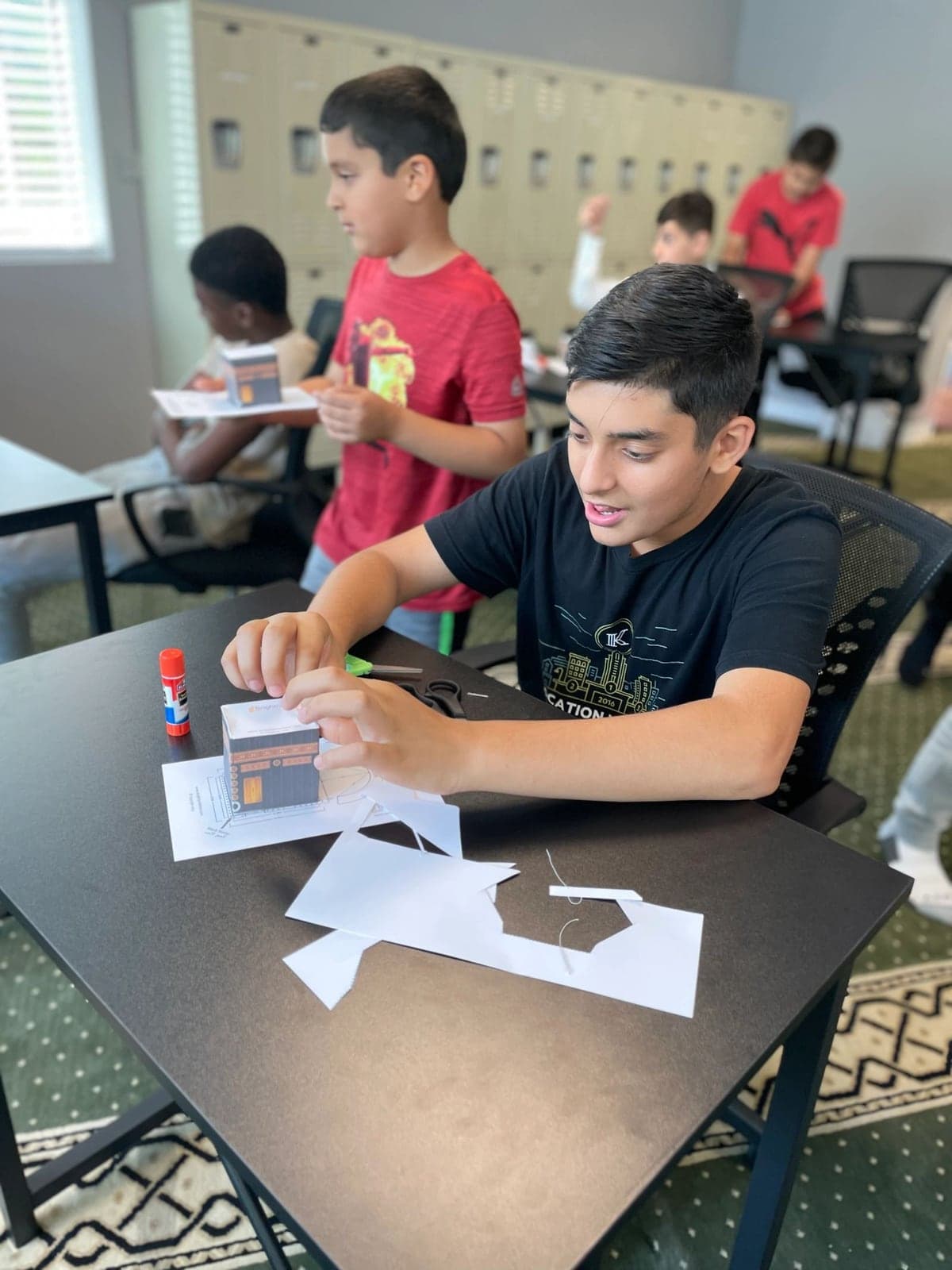 Two boys assembling paper Kaaba models at a workshop table